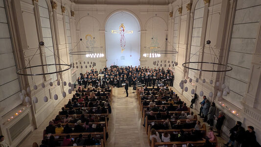 Chor, der auf der Bühne einer Kirche auftritt, während das Publikum in den Kirchenbänken sitzt.