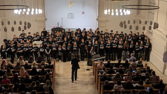 Ein großer Chor singt auf der Bühne einer Kirche vor einem Publikum, das in den Kirchenbänken sitzt.