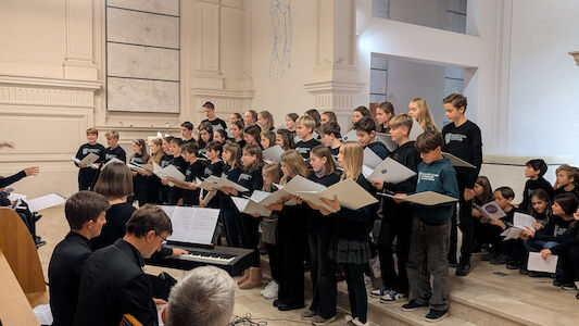 Ein großer Jugendchor singt in einer Kirche, begleitet von einem Pianisten und einem Dirigenten.