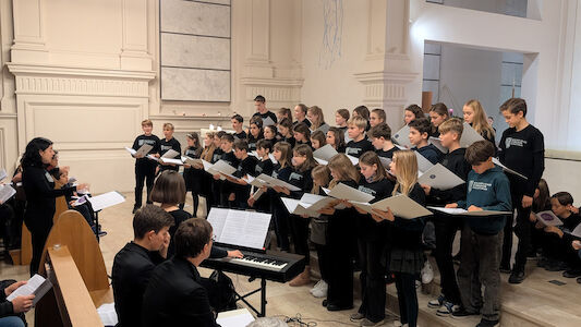 Ein Chor von Kindern und Jugendlichen singt mit einem Pianisten und einem Dirigenten in einem hellen Raum.