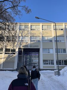 Drei Personen gehen bei strahlend blauem Himmel auf ein schneebedecktes Schulgebäude zu.