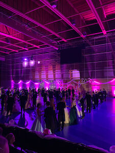 ein Schüler:innenball in einer großen Halle, die Schüler:innen tanzen gerade die Polonaise, der Saal ist in pinkes Licht getaucht