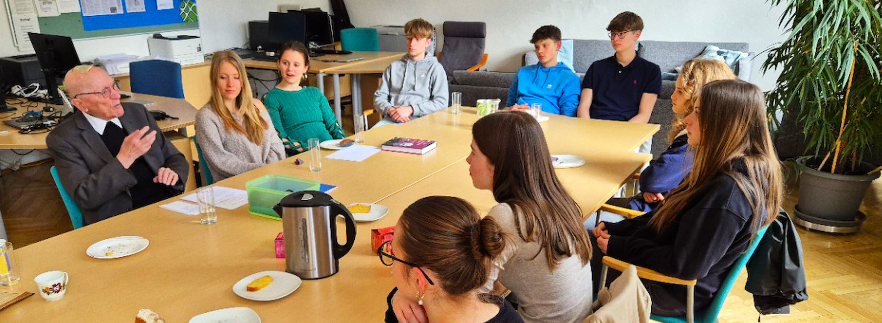 Das Bild zeigt eine Gruppe von Schülerinnen und Schülern, die mit Herrn Generalvikar Städtler an einem langen Tisch in einem modernen, freundlichen Raum sitzen. Der ältere Herr, gekleidet in einen dunklen Anzug, scheint etwas zu erzählen, während die Jugendlichen ihm aufmerksam zuhören.  Auf dem Tisch befinden sich Teller mit Kuchenstücken, Tassen, Gläser mit Wasser sowie eine Edelstahl-Thermoskanne. Einige Teilnehmer haben Blätter und Bücher vor sich. Im Hintergrund sind Schreibtische mit Computern zu sehen, was darauf hindeutet, dass das Treffen in einem Klassenzimmer oder einem Seminarraum stattfindet.  Die Jugendlichen tragen lässige Kleidung, darunter Hoodies, Pullover und T-Shirts. Der Raum wirkt modern und hell mit großen Tischen, Stühlen mit Armlehnen und Pflanzen zur Dekoration. Die Atmosphäre scheint freundlich und konzentriert zu sein, möglicherweise handelt es sich um ein Gespräch zwischen den Schülern und dem älteren Mann über ein wichtiges Thema; eine Zeitzeugenbegegnung.