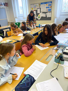 Auf dem Foto ist ein Klassenzimmer mit mehreren Schülerinnen und einer Lehrerin zu sehen. Die Schülerinnen sitzen in Gruppen an zusammengeschobenen Tischen und arbeiten gemeinsam an Arbeitsblättern. Im Vordergrund tauschen sich zwei Mädchen angeregt aus, während sie mit einem Stift Notizen machen. Die Lehrerin steht im Hintergrund und beugt sich über eine Schülerin, um ihr beim Lernen zu helfen.