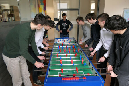 Acht Personen spielen Tischfußball, während eine Person im Hintergrund zusieht.