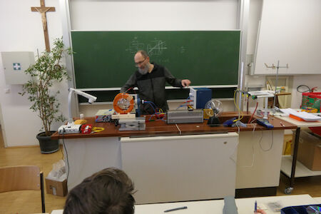 Ein Lehrer demonstriert in einem Klassenzimmer vor einer Tafel ein physikalisches Experiment.