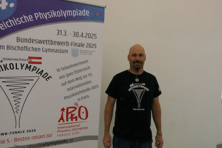 Ein Mann steht neben einem Plakat für die Österreichische Physik-Olympiade 2025.