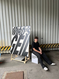 Eine Person in Schwarz sitzt neben einem großen Kunstwerk mit fettgedrucktem Text in einer industriellen Umgebung.