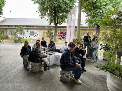 Eine Gruppe von Menschen sitzt und unterhält sich im Freien unter Bäumen in der Nähe eines Gebäudes.