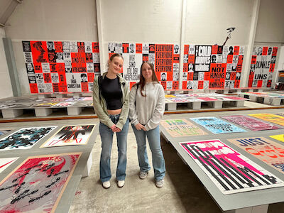 Zwei Frauen stehen in einer Kunstgalerie, in der bunte Poster auf Tischen ausgestellt sind.