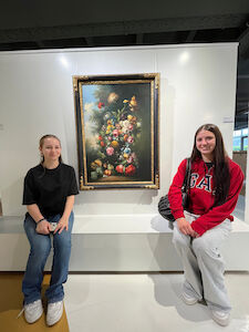 Zwei junge Frauen sitzen neben einem gerahmten Blumenbild in einer Kunstgalerie.