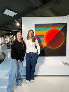 Zwei Menschen stehen lächelnd neben bunter abstrakter Kunst in einer modernen Galerie.