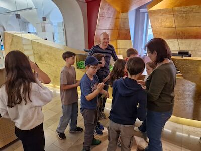 Eine Gruppe von Kindern und zwei Erwachsenen versammelt sich in einem modernen Foyer eines Gebäudes.
