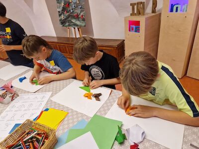 Drei Kinder falten buntes Origami-Papier an einem Tisch mit Kunstzubehör.